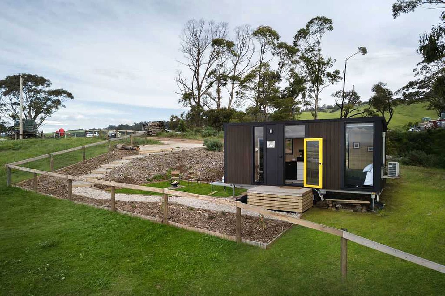 A modern, compact cabin with a glass door, situated on a grassy area with a dirt path leading to it.