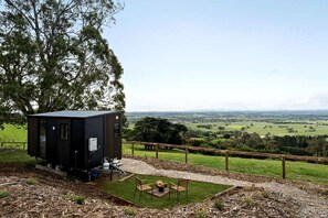 A small, dark-colored cabin with a fire pit and seating area, surrounded by a wooden fence and a grassy field with a view of a distant landscape. - Farm Stay Tiny House with Firepit Under the Stars in Yarragon, Australia (Yarragon South)