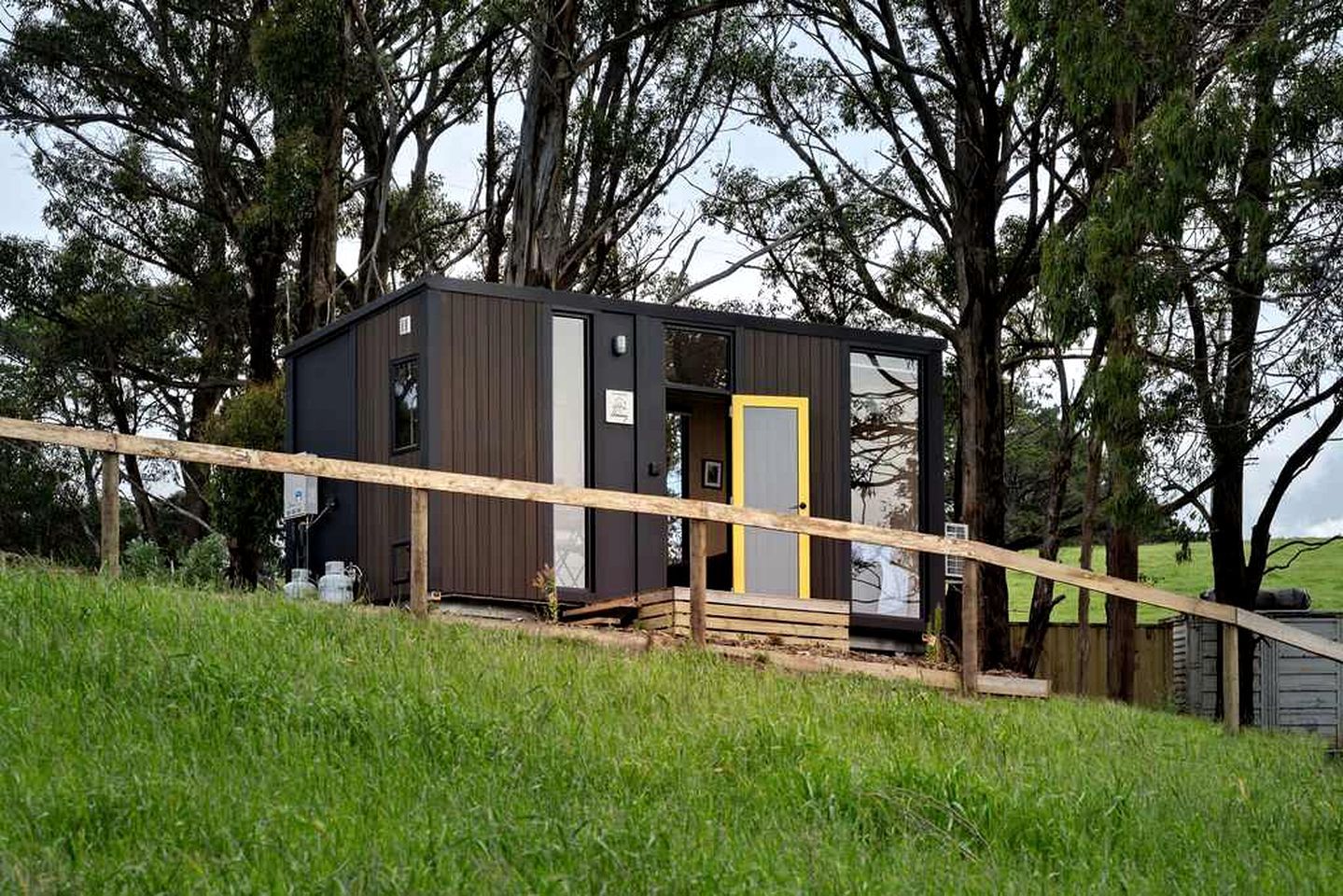 A small, dark-colored cabin with a yellow door, surrounded by trees and a grassy area.