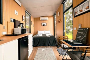 A compact room with a bed, a desk, a chair, a small kitchenette, and a large window. - Farm Stay Tiny House with Firepit Under the Stars in Yarragon, Australia (Yarragon South)