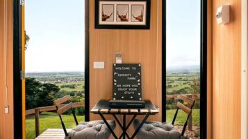Habitación con estructura de madera y vista al campo, un cartel de bienvenida y una mesa con dos sillas acolchadas.
