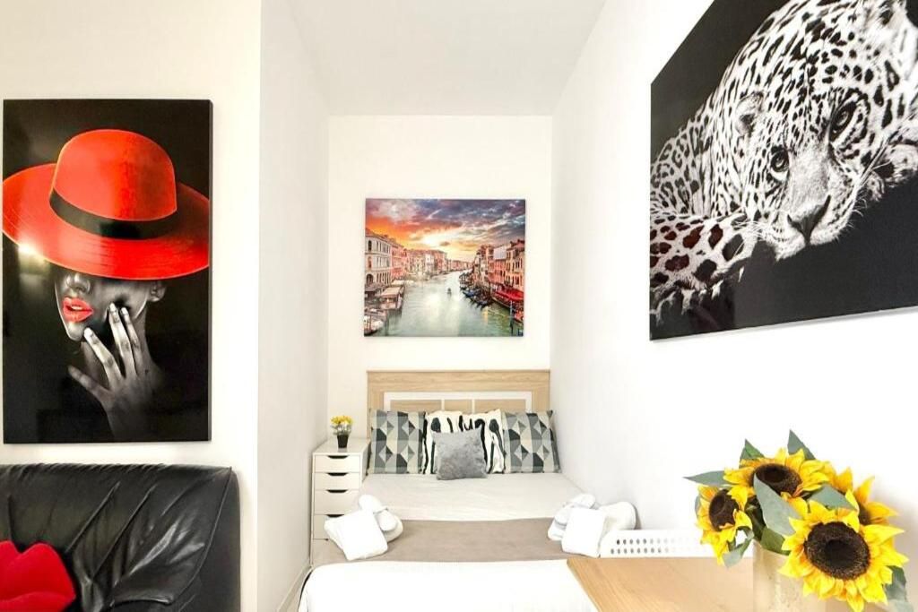 Une chambre d’hôtel avec un lit, une table de chevet, une œuvre d’art représentant un chapeau rouge, un tableau représentant un paysage urbain, une œuvre d’art à motifs léopard et un vase rempli de tournesols.