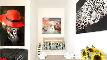 Habitación de hotel con cama, mesita de noche, una obra de arte con un sombrero rojo, un cuadro de paisaje urbano, una obra de arte con estampado de leopardo y un jarrón con girasoles.