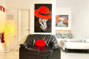Una habitación con un sofá de cuero negro, un cojín con forma de labios rojos, una cama con ropa de cama blanca y dos obras de arte en la pared.