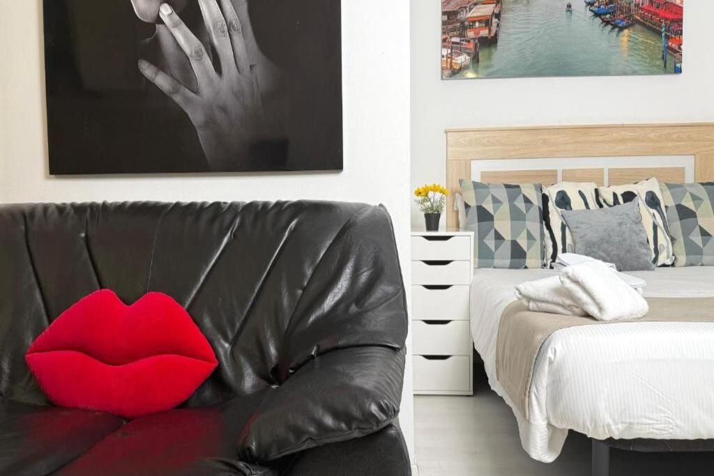 Une pièce avec un canapé en cuir noir, un coussin rouge, un lit recouvert d’une couette blanche et un tableau représentant une main, accroché au mur.