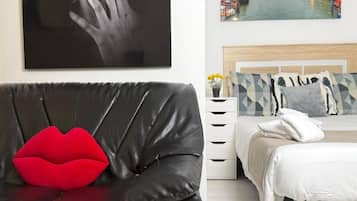 Una habitación con un sofá de cuero negro, un cojín rojo, una cama con edredón blanco y un cuadro colgado en la pared que muestra una mano.
