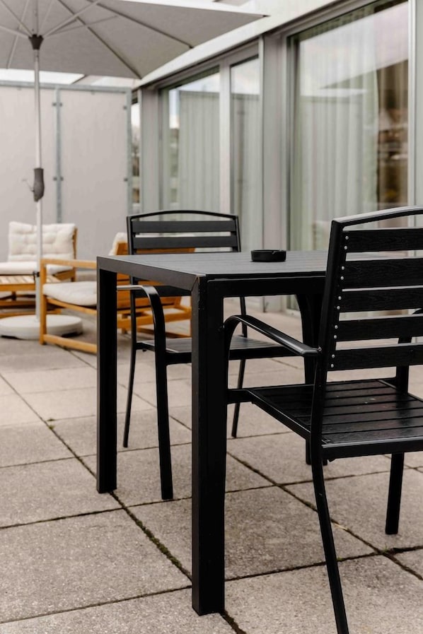 An outdoor patio area with a table and chairs, a ceiling fan, and a view of a building with large windows. - Hine Adon Apart Bern Airport (Belp)