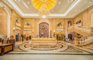 A grand lobby with a central reception desk, a chandelier, and a staircase.