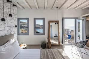 A bedroom with a stone wall, a bed, a large mirror, and a view of the sea. - Saluti Da Stampalia (Astypalaia)