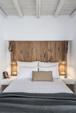 Un dormitorio con cabecera de madera, ropa de cama blanca, dos lámparas de noche y una mesita de noche con un libro y gafas.