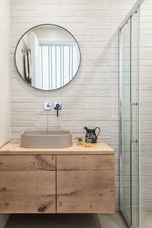 Un baño con un espejo redondo, un lavabo color beige, armarios de madera y una mampara de cristal para la ducha.