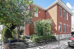 A red brick building with a corner entrance, a small front garden with a tree, and a parked car. - Hannah's Haus Historic German Village 3BR+Parking (Columbus)