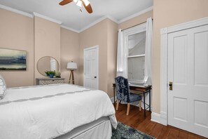 Une chambre à coucher comprenant un lit, un bureau, une chaise, une fenêtre avec des stores, une porte et un tableau accroché au mur.