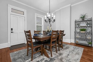A dining room with a wooden table set for four, a chandelier, and a metal shelf with plants and a welcome sign. - Hannah's Haus Historic German Village 3BR+Parking (Columbus)