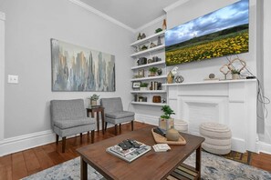 A living room with a fireplace, two grey armchairs, a coffee table with magazines, and a large painting above the fireplace. - Hannah's Haus Historic German Village 3BR+Parking (Columbus)