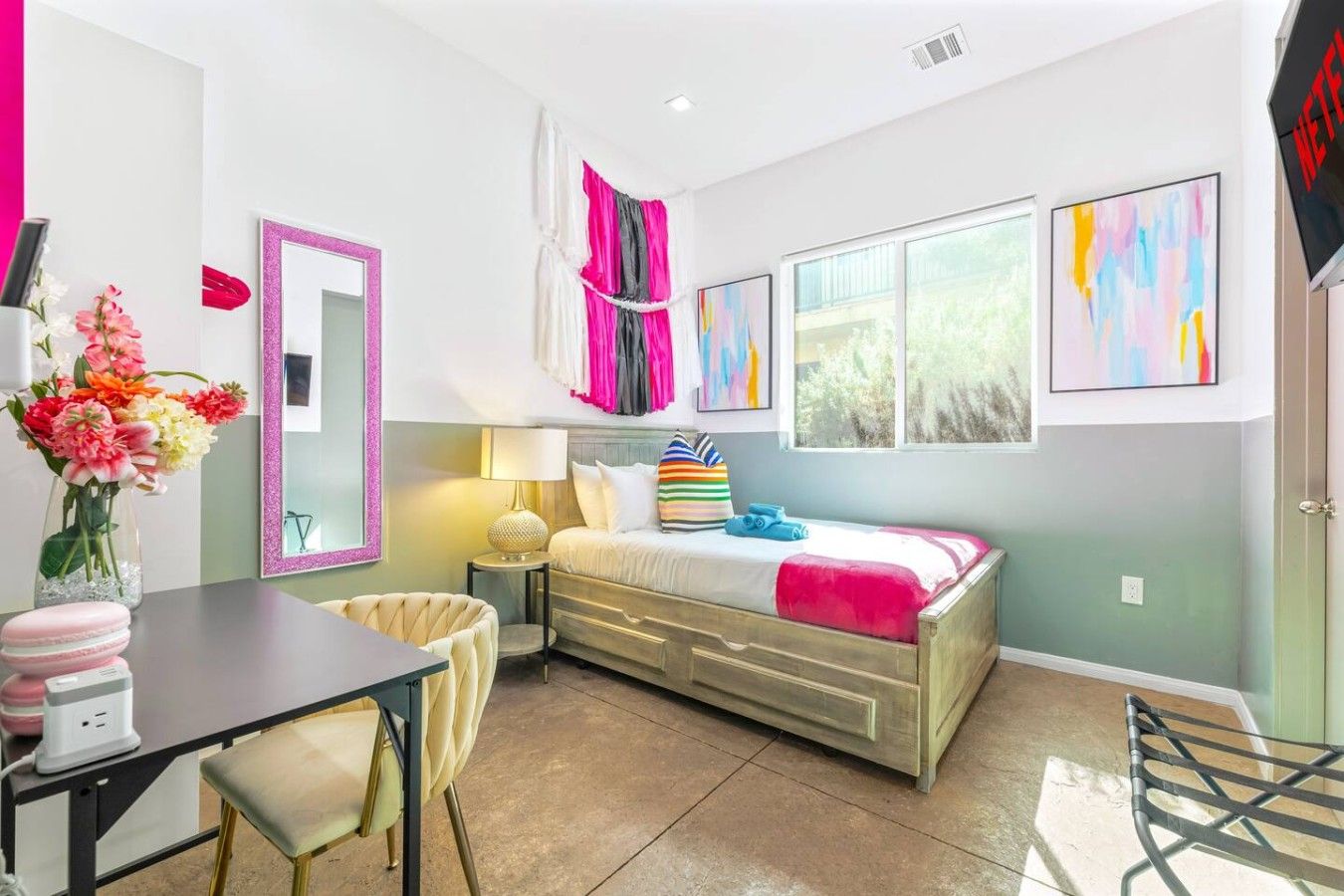 A bedroom with a bed, bedside table, lamp, and a window with artwork.