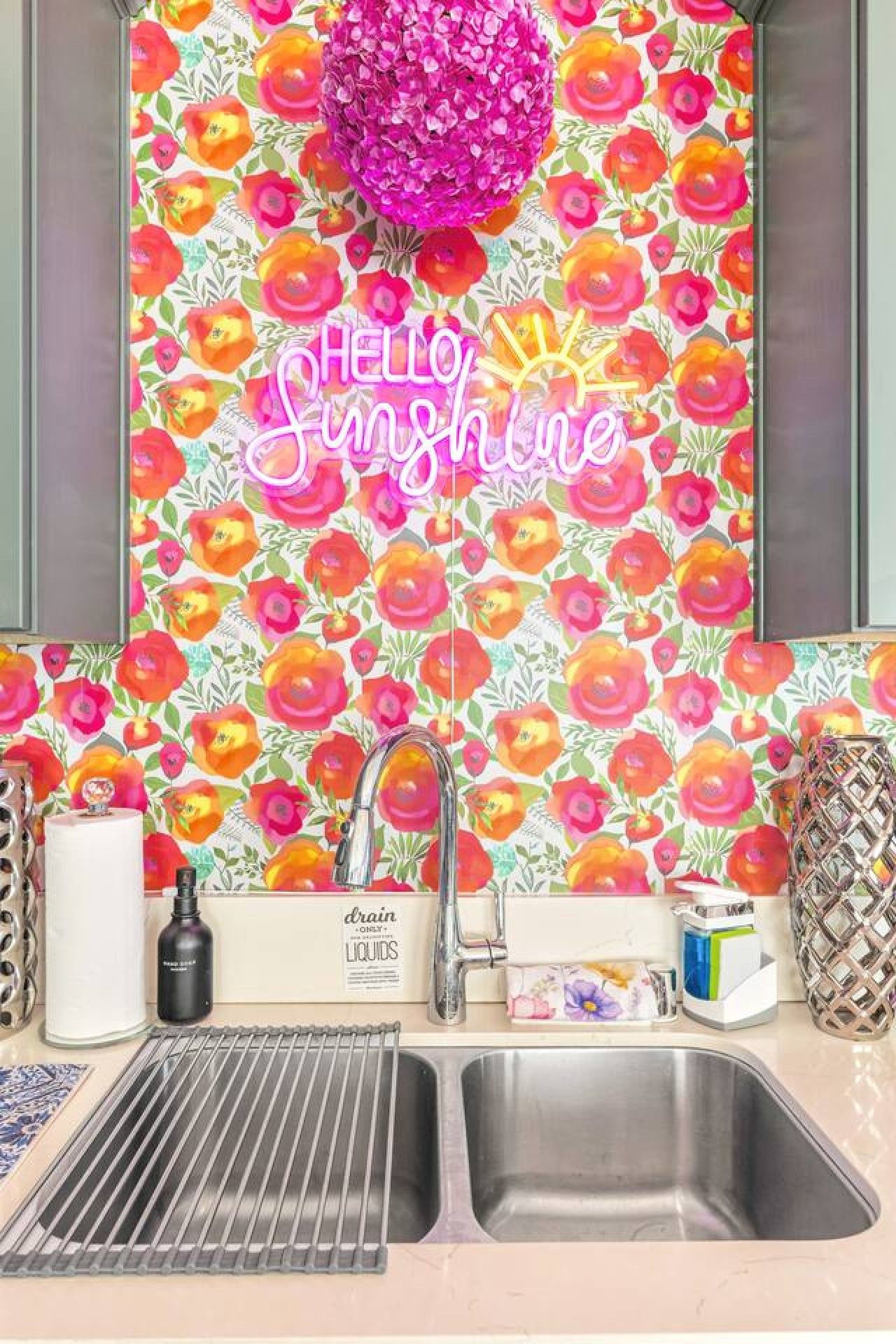 A kitchen with a vibrant floral backsplash, a stainless steel sink, and a 'Hello Sunshine' neon sign.