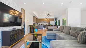 A modern living room with a large flat-screen TV, a sofa, a coffee table, and a dining area with a kitchen in the background.