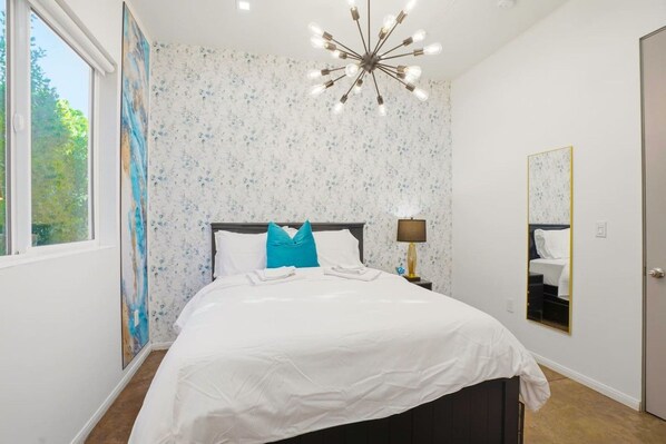 A bedroom with a bed, a nightstand, a lamp, a mirror, and a window.