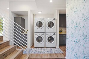 A laundry room with four stacked washing machines, a wooden staircase, and a kitchen area with cabinets.