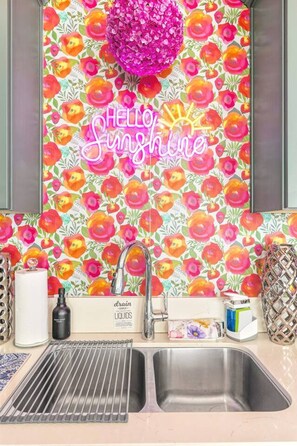 Eine Küche mit einem lebhaften, floralen Rückwand, einem Edelstahl-Spülbecken und einem Neon-Schild mit der Aufschrift 'Hello Sunshine'.
