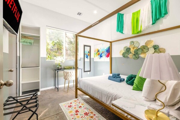 A modern bedroom with a four-poster bed, a desk, and a view of greenery outside.