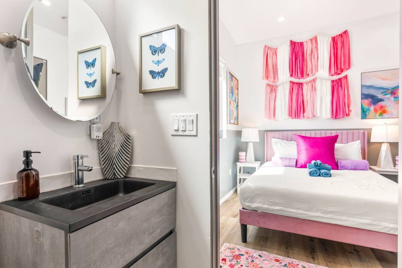 A bathroom with a sink, mirror, and framed butterfly art. The room has a bed with pink bedding and a nightstand with a lamp.