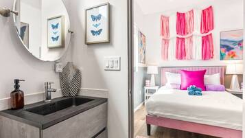 A bathroom with a sink, mirror, and framed butterfly art. The room has a bed with pink bedding and a nightstand with a lamp.