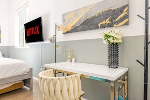 A modern hotel room with a bed, a desk with a vase of flowers, a wall-mounted TV, and a marble artwork.