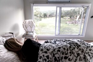 A bedroom with a bed, a bedside table with a lamp, and a window with a view of the outdoors.