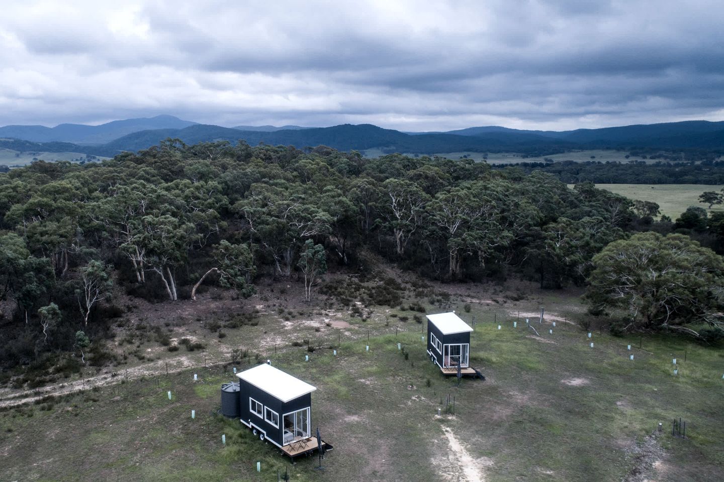 Two small cabins in a grassy field with a forested background and distant hills.