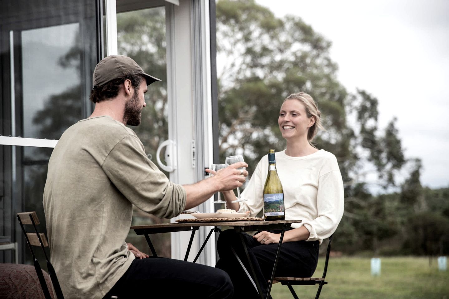 Zwei Personen sitzen an einem Tisch im Freien, mit einer Weinflasche, einem Glas und einem Teller, vor einer Glastür und Bäumen im Hintergrund.