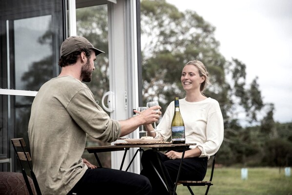 Two people seated at an outdoor table with a bottle of wine, a glass, and a plate, in a setting with a glass door and trees in the background.