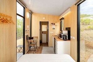 A compact room with a bed, a desk, a chair, a small kitchenette, and a view of the outdoors. - Elevated Tiny House with Valley and Vineyards Views in Victoria (Steels Creek)