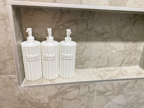 En badeværelseshylde med tre hvide flasker mærket 'Body Wash', 'Conditioner' og 'Shampoo'.