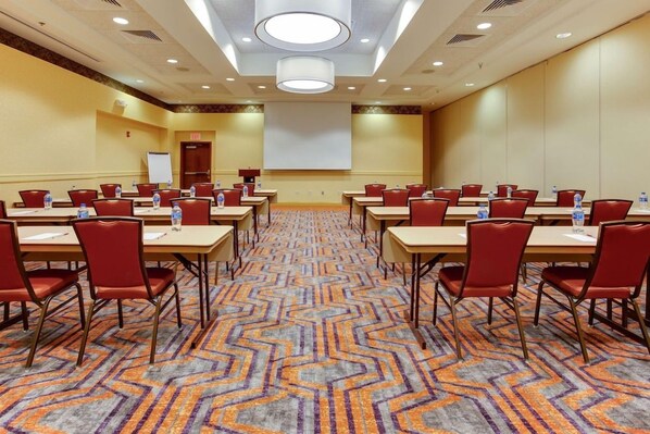 Un salón de conferencias con varias mesas y sillas, piso alfombrado y una pantalla de proyección.
