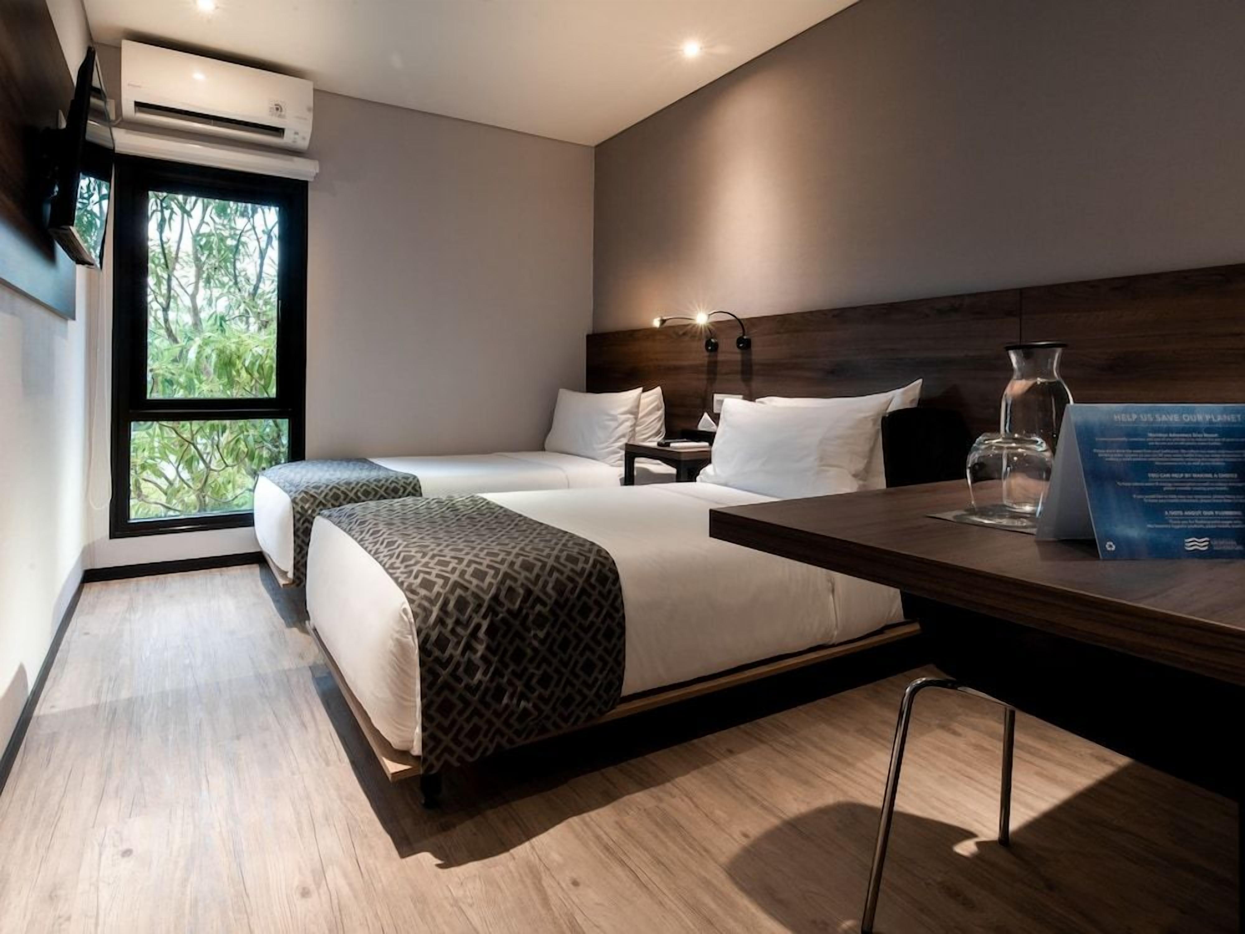 Ein modernes Hotelzimmer mit zwei Betten, Holzboden, einem großen Fenster und einem Schreibtisch mit einem Glas.