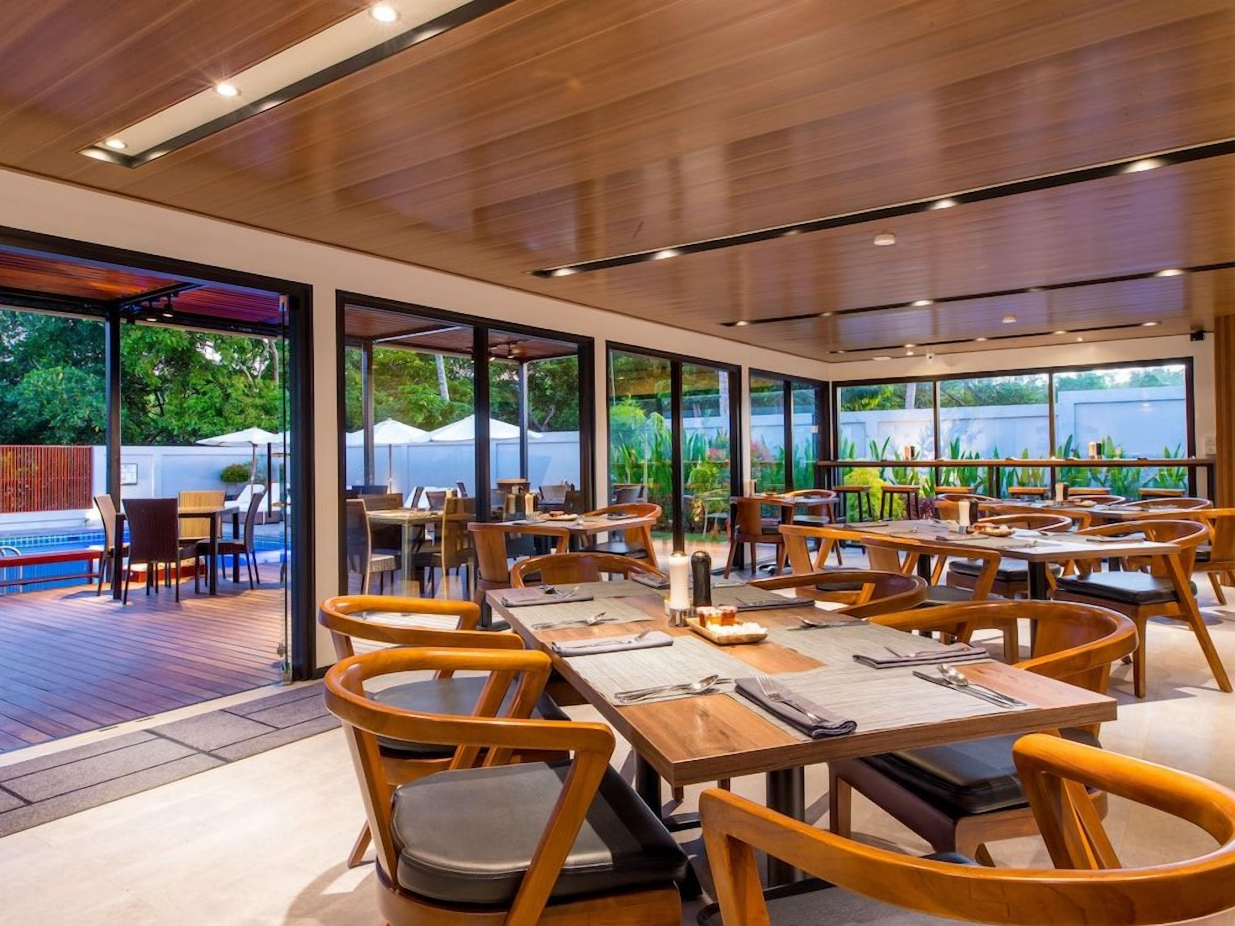 A restaurant with multiple tables and chairs, large windows, and a pool area visible through the glass doors.