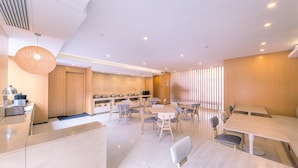 Uma sala de jantar moderna com mesas e cadeiras, cozinha ao fundo e teto com iluminação embutida.