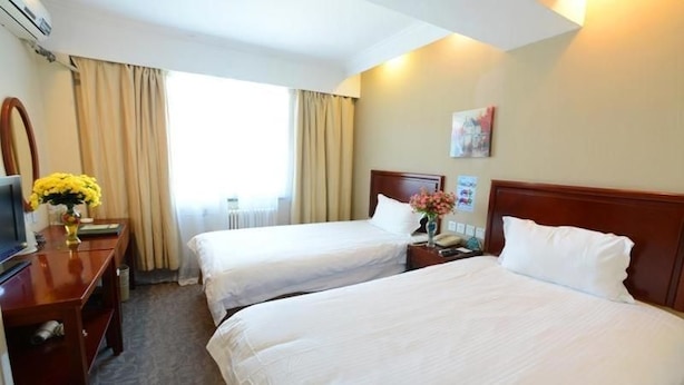 Ein Hotelzimmer mit zwei Betten, einem Schreibtisch mit Computer, einer Vase mit Blumen und einem Fenster mit Vorhängen.