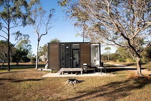 A modern, compact cabin with a sliding door entrance, a small porch with seating, and a barbecue grill outside.