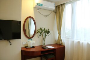 Habitación de hotel con televisión de pantalla plana, escritorio con espejo, planta, ventana con cortinas translúcidas y equipo de aire acondicionado.