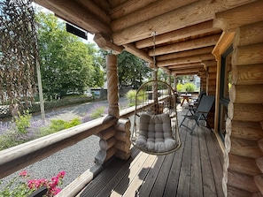 Eine hölzerne Veranda mit einem Hängestuhl und einem Tisch, umgeben von viel Grün und einem Kiesweg.