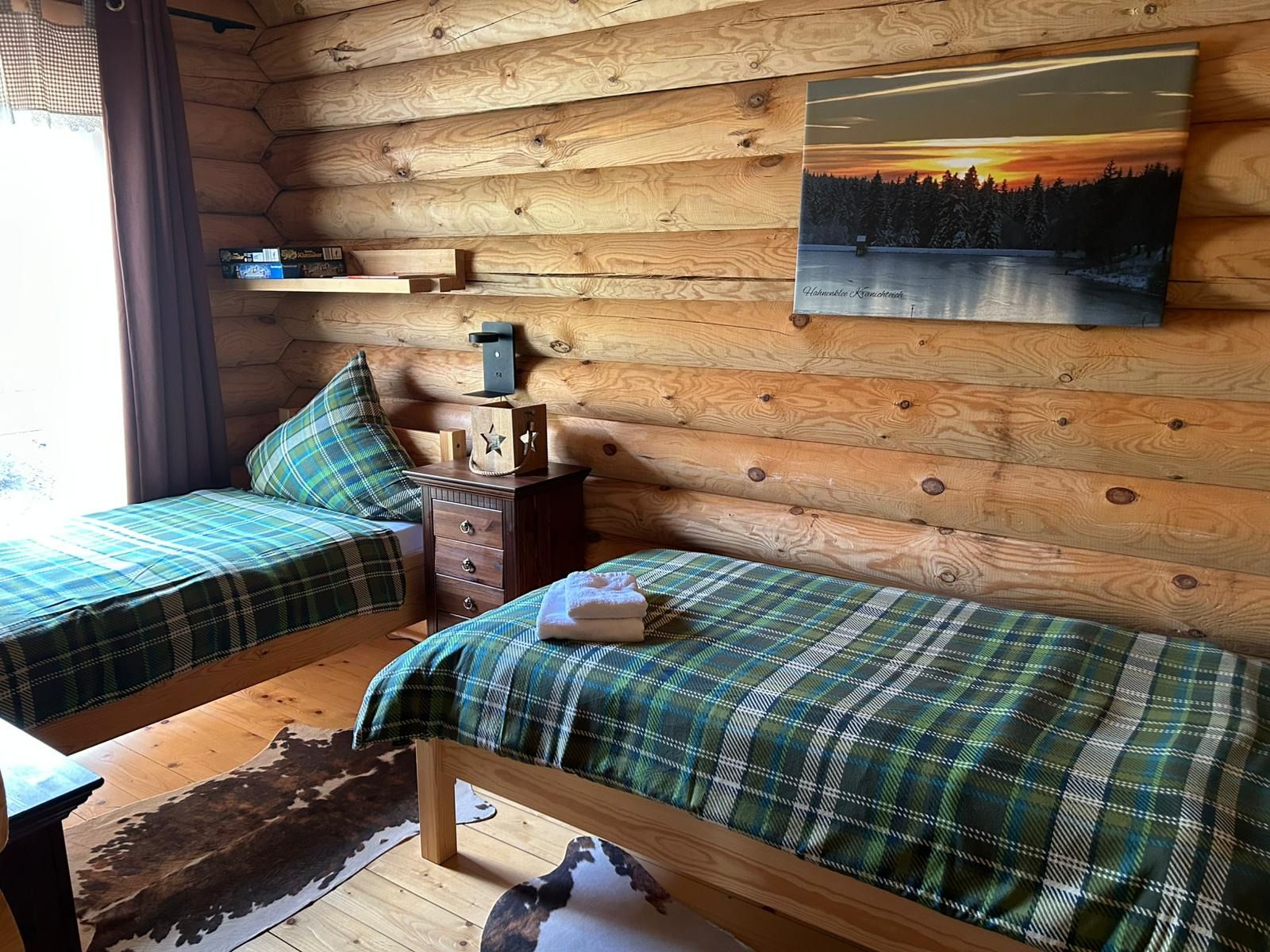 Ein Blockhauszimmer mit zwei Betten, einem Holzkleiderschrank, einem Rindsfellteppich und einem Sonnenuntergangsbild.