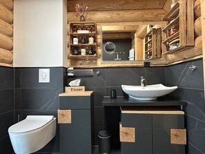 Un baño moderno con inodoro blanco, lavamanos con espejo arriba y una caja de pañuelos con la etiqueta 'Tissue' sobre una encimera de madera.
