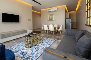 A modern living room with a grey sofa, a coffee table, and a dining area with white chairs. - Mirada Gold - Obhur (Jeddah)