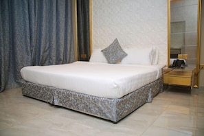 A neatly made bed with a white mattress and a patterned bed skirt, a bedside table with a telephone, and a mirror on the wall. - Mirada Gold - Obhur (Jeddah)