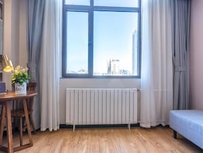 A room with a large window, a radiator, a wooden floor, a table with a lamp, and a chair.
