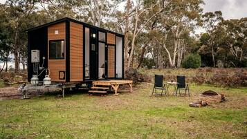 Une petite cabane au revêtement extérieur en bois, avec un banc, deux chaises et une table, située dans un espace verdoyant, avec des arbres en arrière-plan.