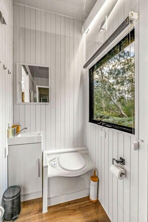 A modern bathroom with a white sink, toilet, and a window with a view of trees. - Cozy Tiny House with Barbecue, Firepit and River Views in Victoria, Australia (Dadswells Bridge)